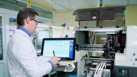 The engineer in a white robe and glasses works for the Surface Mount Technology machine. PCB Processing on CNC machine, Production of electronic components at high-tech factory