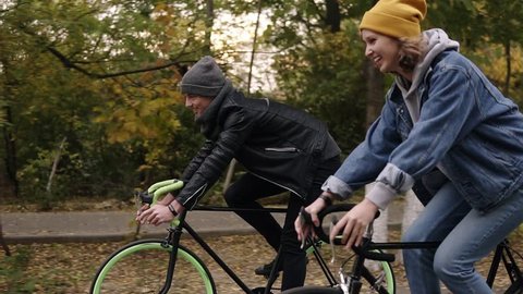 Smiling, young hipster couple enjoying cycling through park on trekking bikes. Two young people in hats having great time together in autumn. Side view. Slow motion