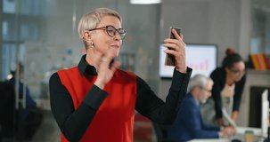 Mature caucasian businesswoman making video call using her mobile phone and talking to her friends standing in modern office