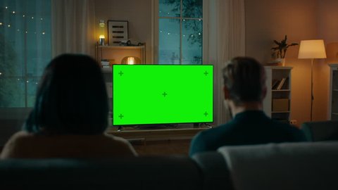 Couple Watches Green Mock-up Screen TV while Sitting on a Couch in the Living Room. Romantic Evening for Boyfriend and Girlfriend.