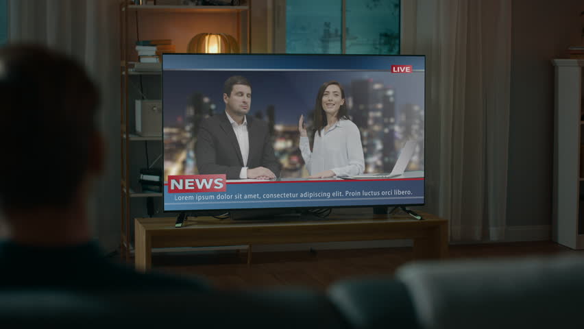 Man Watches Late Night News Show on TV while Sitting on a Couch at Home in the Evening. Two Presenters Talk and Joke on TV. Cozy Living Room with Warm Lights. Over the Shoulder Shot.