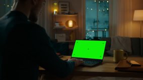 Man Sitting at His Desk Works on a Laptop with Green Chroma Key Screen. Late at Night in His Living Room Man Uses Notebook Computer. Side View Zooming on Screen.