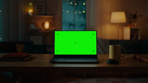 Laptop Computer Showing Green Chroma Key Screen Stands on a Desk in the Living Room. In the Background Cozy Living Room in the Evening with Warm Lights on. Zoom In Shot.