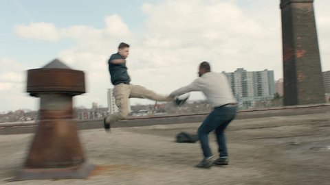 Angry and quick skinny man runs across a warehouse rooftop and attacks a buff man with skilled kicks and punches in overcast sunlight. Medium shot in 4K with an Alexa Mini camera