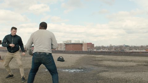 Two fit men fight on a warehouse rooftop with skilled kicks and punches in overcast sunlight. Medium shot in 4K with an Alexa Mini camera