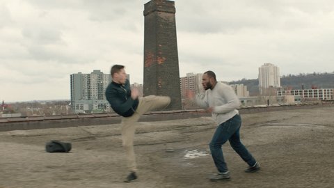 Two men fight on a warehouse rooftop with skilled kicks and punches in overcast sunlight. Medium shot in 4K with an Alexa Mini camera