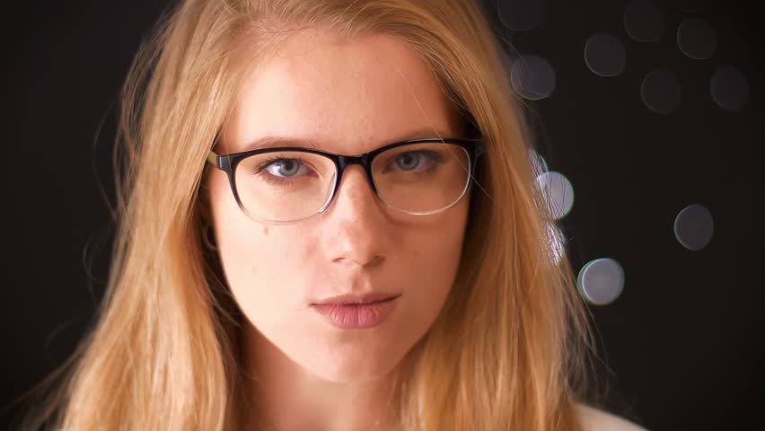 Relaxed blonde caucasian girl in glasses with smart look , touching her glasses confidently while looking at camera with artistic face, dark studio illustration