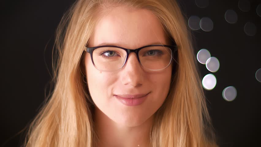 Confident caucasian blonde woman is looking precisely in her glasses at camera and smirking with calm face on dark background