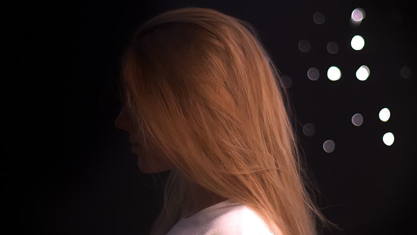 Cute caucasian woman with long blonde hair is turning her body and looking at camera precisely through glasses, while standing isolated on black background