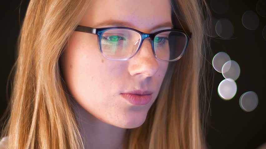 Reflection of green screen in spectacles on caucasian blonde female, who is standing focused on using device and being relaed on dakr background