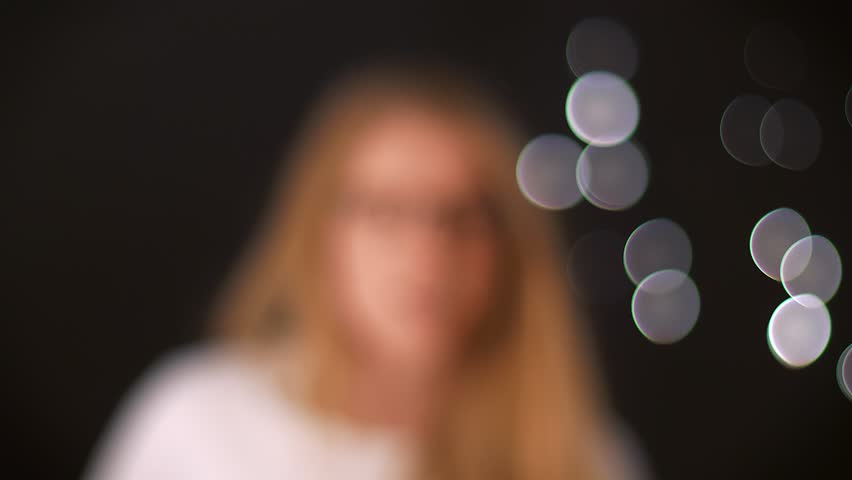 Shooting spectacled girl goes towards camera from blurred black background, being focused, happy gorgeous face