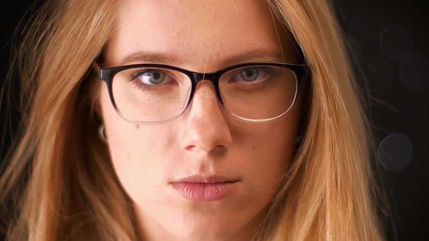 Portrait of nice blonde spectacled caucasian woman looking in camera closely while standing on black background indoor, blurred illustration