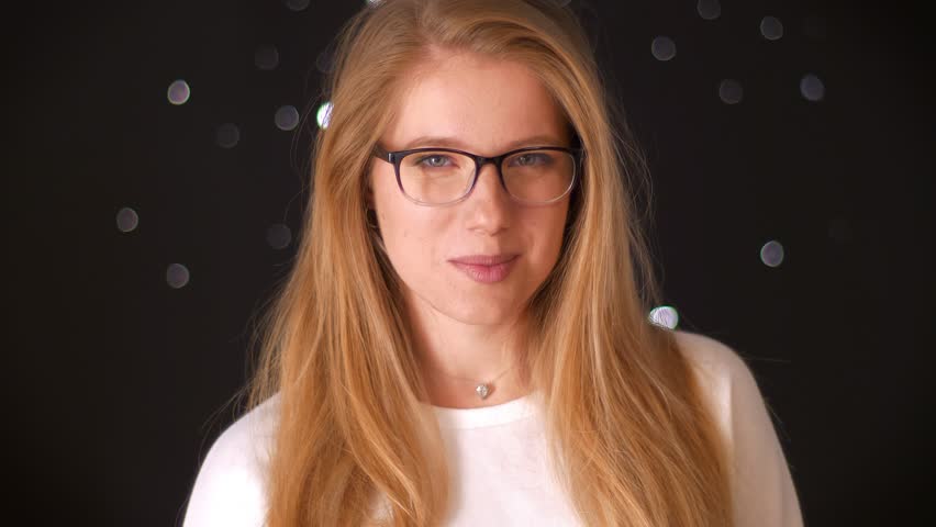Pretty smiling caucasian female is looking at camera and smiling, blonde long hair and glasses on her nice face, chilling on black background