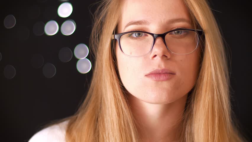 Cute caucasian blonde woman in spectacles is standing on black background and looking at camera with assured face, indoor