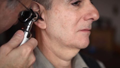 A doctor examines a senior man's ear with an otoscope in a home environment.