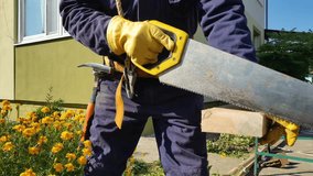 Man in protective gloves and blue safety helmet work with hand saw outdoors. Clear confident movements professional carpenter sawing a wooden beam, close-up, manual labor stock video