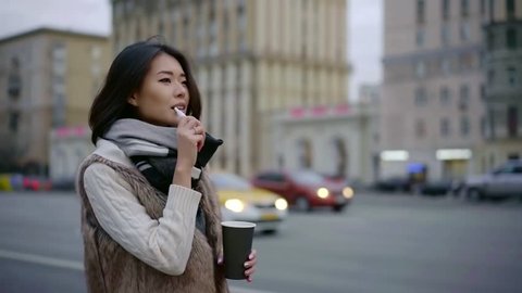 confident asian woman is vaping and drinking coffee walking on city street, moving near road