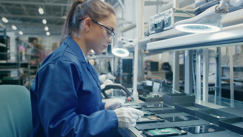 Woman Electronics Factory Worker in Blue Work Coat and Protective Glasses is Assembling Smartphones with Tweezers and Screwdriver. High Tech Factory Facility with more Employees in the Background.  Royalty-Free Stock Footage #1020186892