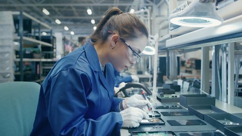 Woman Electronics Factory Worker in Blue Work Coat and Protective Glasses is Assembling Smartphones with Tweezers and Screwdriver. High Tech Factory Facility with more Employees in the Background. 