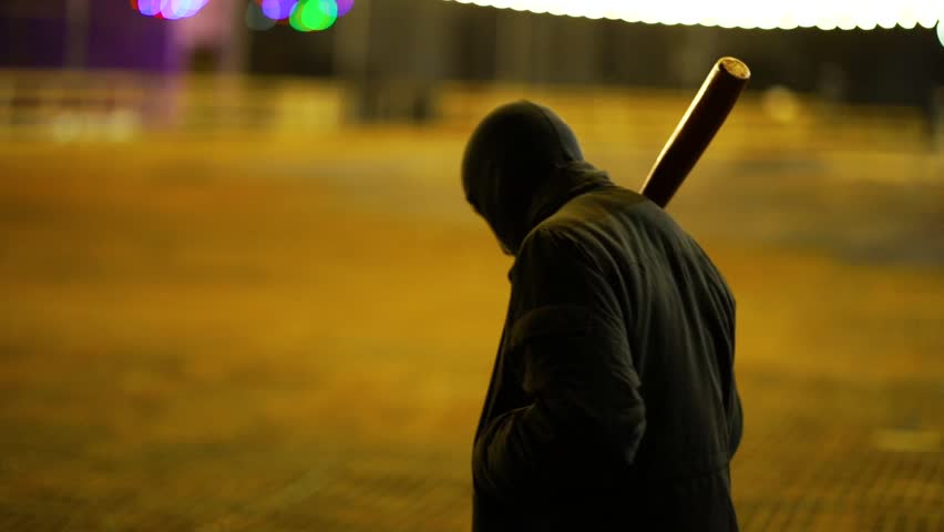 A man dressed in dark clothes and a balaclava goes with a bat in an abandoned park. There is an early winter outside and garlands hang around. His face is not visible in backlight. He is set up to fig