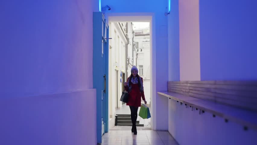 Young white woman in winter clothes is walking in the corridor of the office or shopping center. Smiling girl has shopping bags in her hands. Hallway is modern and neon lightened. 