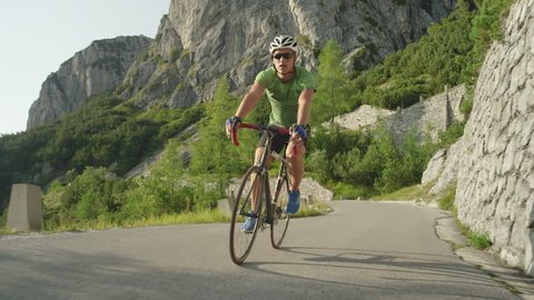 SLOW MOTION, LOW ANGLE: Male road biker speeding down an asphalt road in the scenic mountains in Slovenia. Active Caucasian man enjoying the summer by road biking in the beautiful Slovenian nature.