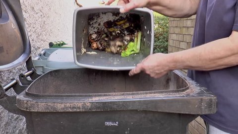 POV close shot of a man's hands raising the lid of a large waste bin and pouring in non-recyclable food waste from a smaller bin.