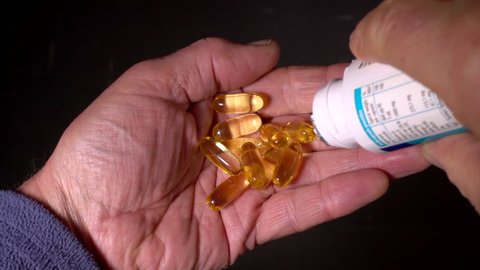 Slow motion close POV shot of cod liver oil capsules (rich in vitamins A and D) being poured from a small plastic bottle / container into the palm of an elderly / senior man's hand.