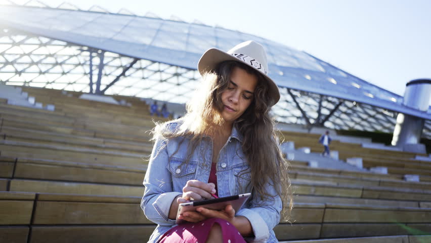 Panning aroung girl using tablet. Portrait Young casual woman sitting on wood tribune in sunshine