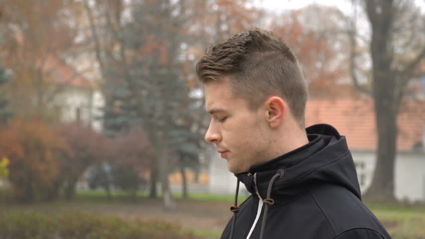 A young man smokes a cigarette and lets go of smoke outside on a cold autumn day.