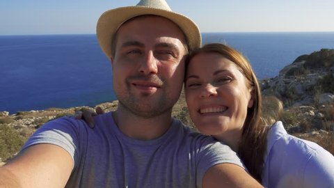 POV view of funny loving young couple doing selfie at edge of cliff. Girlfriend kissing boyfriend on cheek. Breathtaking view of blue Mediterranean sea on background. Concept of romance and travel