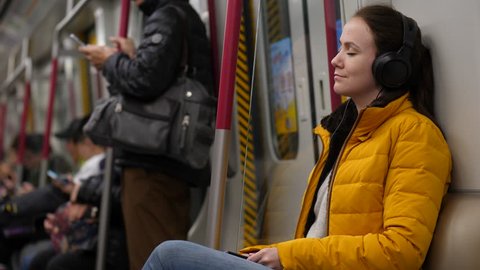 Young adult woman sit with eyes closed in metro train, listen music, large earphones on head. Blurred background, subway car ride between stations. Tired but satisfied person rest at public transport