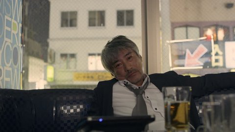Drunk sad Japanese man sitting at a booth in a karaoke room alone with soft interior lighting. Medium shot on 4k RED camera.
