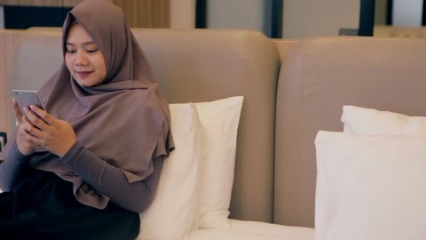 Young asian muslim woman using smartphone on bed and smiling while chatting with friends.