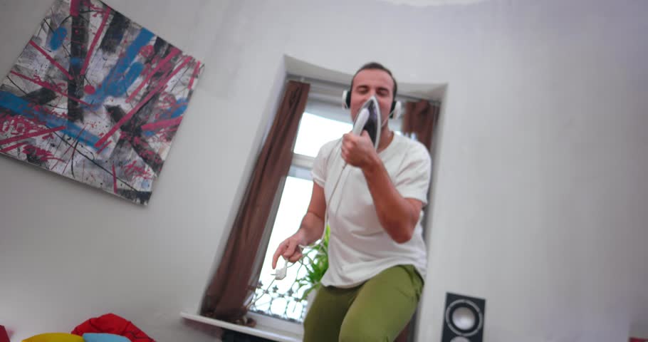 Camera shoots from bottom up. Cheerful caucasian guy looks at camera, listens music in headphones and sings song using an iron as microphone