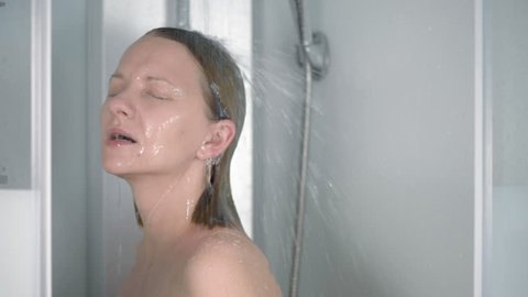 Young woman taking a shower. Beautiful girl washes hair in the white shower cabin at bathroom. Happy adult relaxing the water drops on her body. Slow motion.