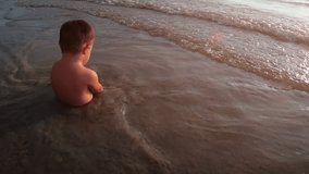 Adorable toddler sits in the shallow water. catching the tiny waves at this tropical beach paradise in Thailand. FullHD footage