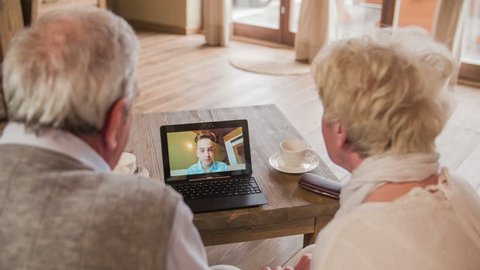 Boy waving over video call with grandparents HD. Over shoulder view of elderly couple calling grandson with video call on laptop computer. Laptop on wooden desk. Two person sit on sofa.