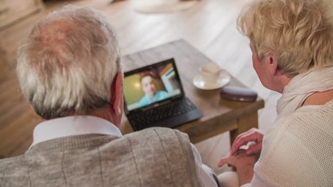 Grandparents talk to grandson over laptop video call HD. Jib shot behind elderly couple sit on sofa with laptop computer on table and having a conversation with boy on screen. Live view skype chat.