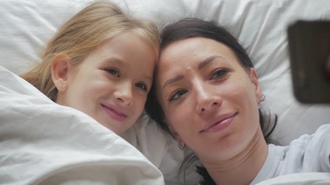 Young beautiful mother and her cute daughter taking selfie with smartphone and smiling while lying on the bed at home. Woman taking selfie with a little girl in bedroom.