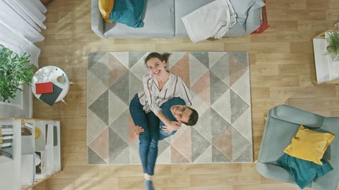Young Happy Couple Looking Above. Man is Holding the Girl in His Arms. Cozy Living Room with Modern Interior with Carpet, Sofa, Chair, Coffe Table, Book Shelf, Plant and Wooden Floor. Top View. 