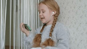 happy little girl with pigtails in headphones looking at the phone video, smiling, sitting in the room on the windowsill, covering herself with a rug, portrait 4k