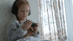 happy little girl with pigtails in headphones looking at the phone video, smiling, sitting in the room on the windowsill, covering herself with a rug, portrait 4k