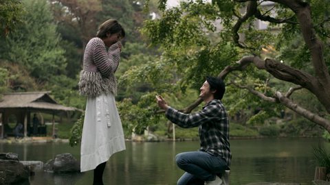 Cheerful Japanese man proposes to his girlfriend and they hug happily in a beautiful garden. Romantic proposal scene. Wide shot on 4k RED camera. 