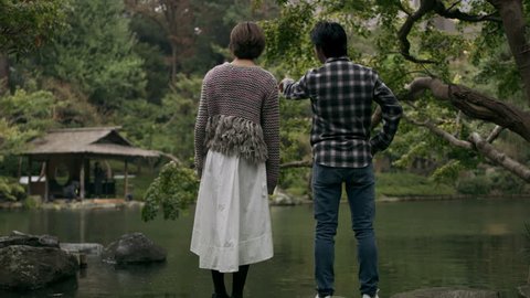 Nervous Japanese man looks out across a pond with his girlfriend and proposes to her in a beautiful garden in the rain with soft natural lighting. Wide shot on 4k RED camera. 