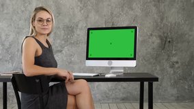 Cheerful young woman sitting in office talking to camera and showing on display of computer something. Green Screen Mock-up Display.