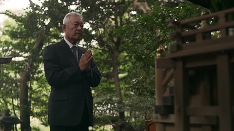 Japanese senior citizen in suit clapping and bowing in front of a shrine in a beautiful green garden with soft natural lighting. Medium shot on 4k RED camera. 