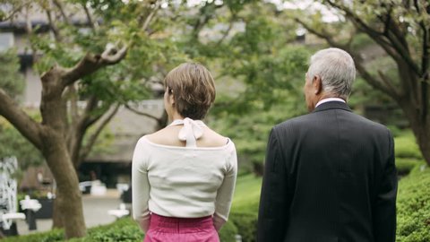 Young adult woman walking and talking with an elderly man along a path in a vibrant garden with soft natural lighting. Medium shot on 4k RED camera. Stockvideó