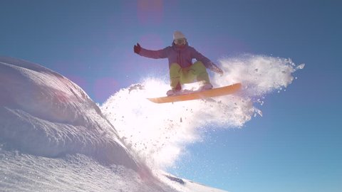 SLOW MOTIONWARP, LENS FLARE, CLOSE UP: Pro snowboarder jumping in fresh snow, spraying snowflakes over sun. Male freerider takes off in the air on his snowboard on sunny day on the mountain slopes.