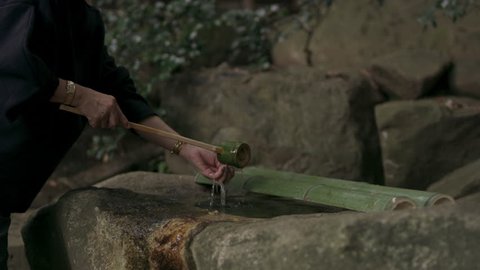 Elderly Japanese woman taking water from a stone water basin and pouring it onto her hands in a beautiful garden with soft natural lighting. Medium shot on 4k RED camera.